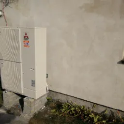 Zewnętrzna jednostka pompy ciepła Mitsubishi Electric ZUBADAN, zamontowana na ceglanych postumentach przy ścianie budynku. Widoczny fragment okna i instalacja na ścianie.