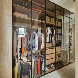 Duża, przeszklona szafa z drewnianymi elementami, wypełniona ubraniami, butami i pudełkami, z widoczną refleksją okna w szklanych drzwiach.