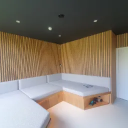 Wnętrze pokoju z narożną, tapicerowaną sofą z drewnianą podstawą, wbudowaną w ścianę z drewnianych lameli. Pod sofą widoczna przestrzeń do przechowywania z akcesoriami do ćwiczeń.