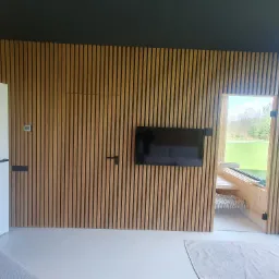 Nowoczesne wnętrze z drewnianą ścianą z listew, ukrywającą drzwi i z wbudowanym telewizorem, z widokiem na zewnątrz przez okno z siedziskiem.