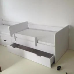 Białe łóżko dziecięce z wysuwaną szufladą i barierką ochronną, widoczne z boku, z jednym butem dziecięcym na podłodze w rogu. Minimalistyczny design.