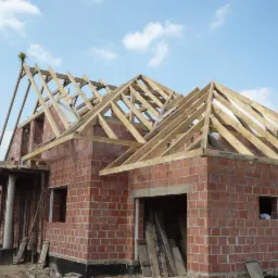 Murowany dom w trakcie budowy, z widoczną konstrukcją drewnianą dachu i częściowo wzniesionymi ścianami z cegły. Niebo z lekkimi chmurami w tle.