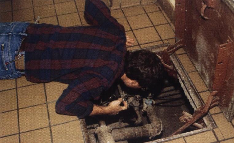 Mężczyzna pochylony nad otwartym włazem w podłodze, prawdopodobnie inspekcja lub naprawa rur w przestrzeni podpodłogowej, widoczne rury i narzędzia.