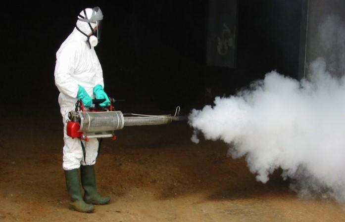 Osoba w kombinezonie ochronnym rozpyla gęstą mgłę za pomocą specjalistycznego urządzenia. Praca w słabo oświetlonym pomieszczeniu.