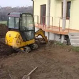 Żółta mini koparka przy budynku z drewnianym tarasem, wykop w ziemi, łopata leży na ziemi, drzewa w tle.