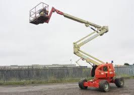 Podnośnik koszowy teleskopowy z czerwoną podstawą i beżowym ramieniem, na nierównym terenie, z widocznym koszem i operatorem na tle pochmurnego nieba.