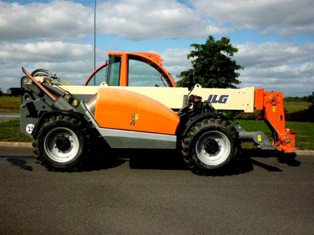 Pomarańczowo-szara koparka teleskopowa JLG z wysięgnikiem na tle pochmurnego nieba i zieleni. Maszyna stoi na asfalcie. Widok z boku, z bliska.