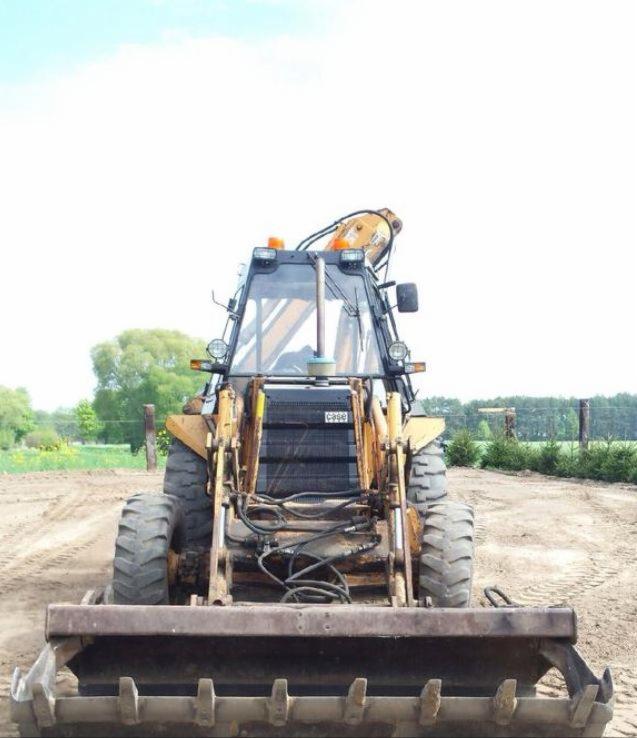 Żółto-czarna koparko-ładowarka Case z przodu, na piaszczystym terenie. Widoczne opony, kabina z lampami i przednia łyżka z zębami. W tle drzewa i niebo.