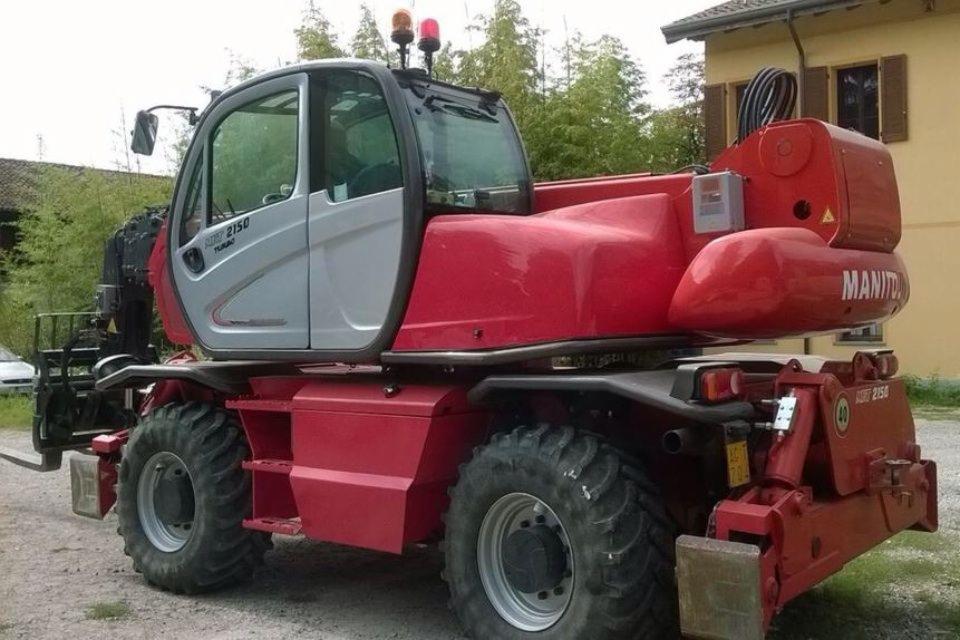 Czerwono-szara koparka Manitou MRT 2150 Turbo z wysięgnikiem teleskopowym na tle zieleni i budynku. Widoczny tył maszyny z dużymi oponami.