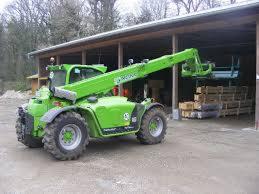 Zielona ładowarka teleskopowa Merlo z podnośnikiem koszowym, zaparkowana przed otwartym magazynem z paletami. Widoczne drzewa w tle, pochmurny dzień.