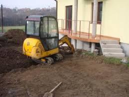 Żółta mini koparka przy budynku z drewnianym tarasem, wykop w ziemi, łopata leży na ziemi, drzewa w tle.