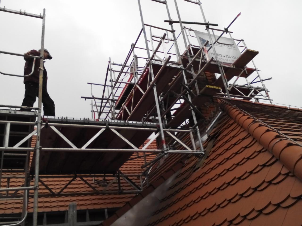 Pracownik na rusztowaniu przy kominie z cegły na dachu pokrytym czerwoną dachówką, podczas prac remontowych. Widok z dołu, pochmurne niebo w tle.