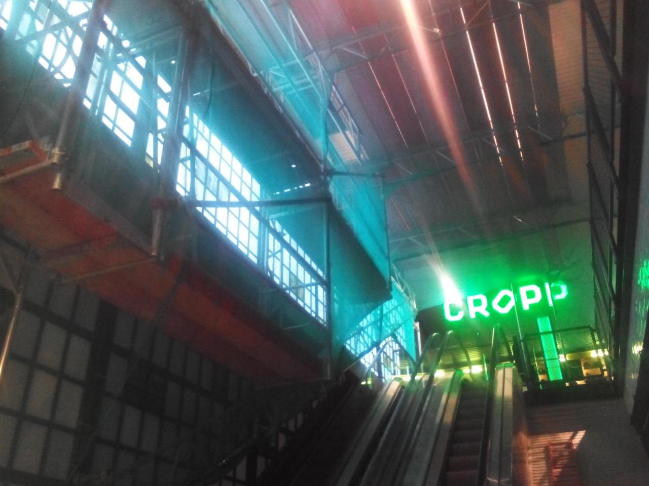 Montaż rusztowania przy schodach ruchomych wewnątrz budynku, widoczny neon CROPP, zabezpieczenia siatką. Prace na wysokościach.