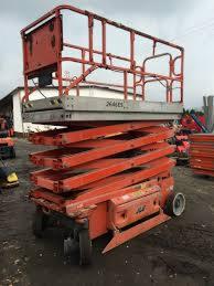 Czerwony podnośnik nożycowy na nierównym terenie; widoczne ślady użytkowania, częściowo widoczne budynki w tle, sprzęt budowlany.