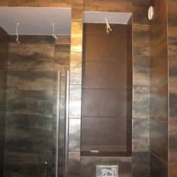 Wykończona łazienka z brązowymi płytkami imitującymi metal, kabina prysznicowa ze srebrnymi ramami i wnęka w ścianie z oświetleniem. Świeżo położona glazura.