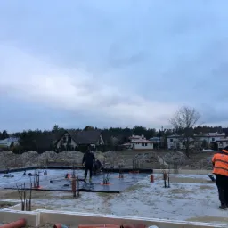 Przygotowanie fundamentów pod budowę domu: robotnicy pracują na izolacji przeciwwilgociowej, widoczne zbrojenia i szalunki styropianowe, w tle zabudowa jednorodzinna.