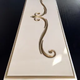 Front szuflady lub drzwiczek meblowych w kolorze kremowym z ozdobnym, złotym ornamentem w stylu retro, leżący na ciemnym blacie.