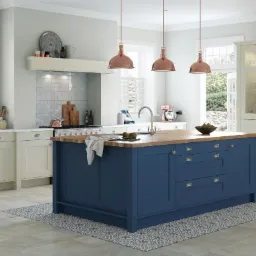 Elegancka, przestronna kuchnia z wyspą w odcieniach bieli i granatu, oświetlona miedzianymi lampami wiszącymi. W tle widać okno z roletami i ogród. Realizacja z Brzegu.