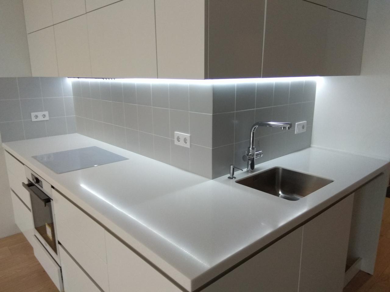 Nowoczesna kuchnia na wymiar z białymi frontami, szarymi płytkami i oświetleniem LED pod szafkami, widoczny zlew ze stali nierdzewnej i płyta indukcyjna.