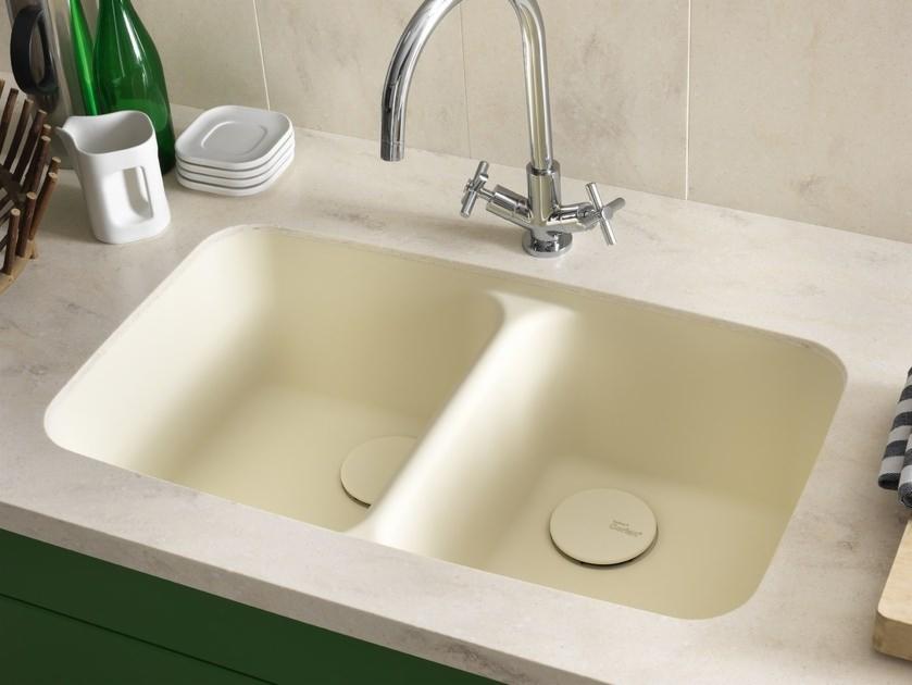 Beżowy dwukomorowy zlew z chromowaną baterią na blacie z zielonymi szafkami, widoczne akcesoria kuchenne i deska do krojenia.