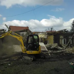 Żółta mini koparka JCB 8025 w trakcie rozbiórki drewnianego budynku z żółtymi elementami, widoczne unoszące się pyły i gruz, taczka z narzędziami w prawym dolnym rogu.