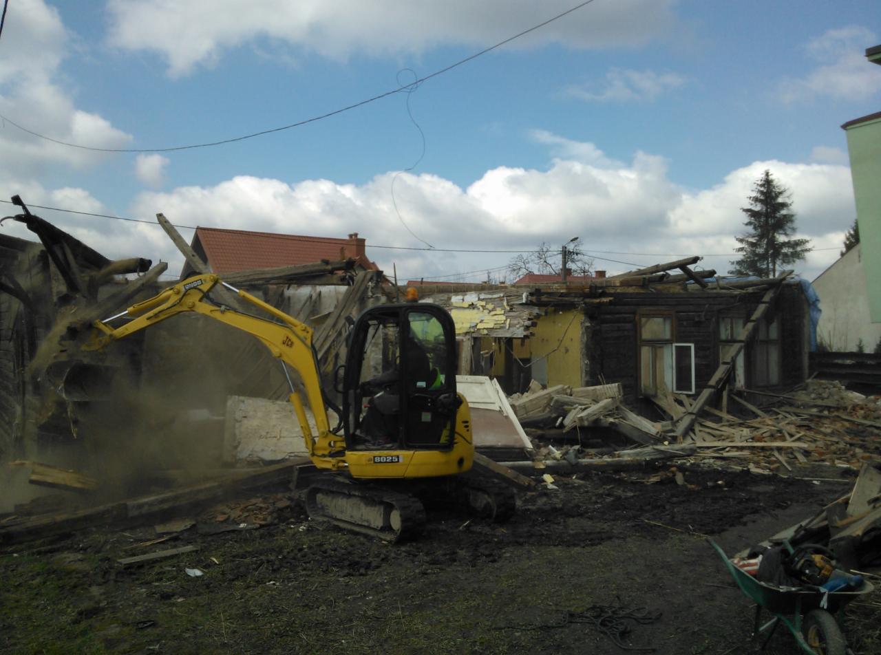 Żółta mini koparka JCB 8025 w trakcie rozbiórki drewnianego budynku z żółtymi elementami, widoczne unoszące się pyły i gruz, taczka z narzędziami w prawym dolnym rogu.