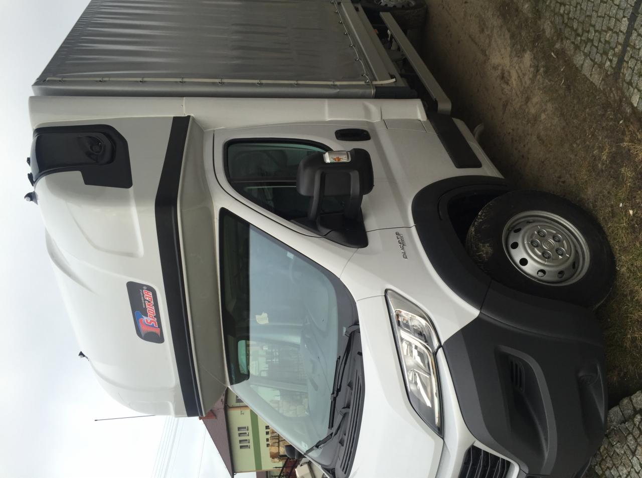 Biały bus z plandeką marki Fiat Ducato, zaparkowany na poboczu, z widocznym logo 'Spokar' na kabinie. Ujęcie z bliska, lekko z dołu, z widocznym fragmentem budynku w tle.