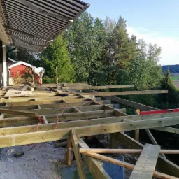 Konstrukcja drewnianego tarasu w trakcie budowy; widoczny szkielet z belek, betonowe podpory, narzędzia pracy oraz pracownik w oddali. W tle zieleń drzew i budynek z markizą.