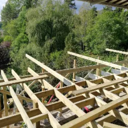 Drewniana konstrukcja tarasu w trakcie budowy, widok z góry na szkielet z belek i metalowych łączników na tle zieleni drzew.
