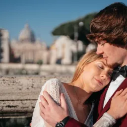 Para młoda w objęciach na tle rzymskiej architektury, panna młoda w koronkowej sukni z zamkniętymi oczami, pan młody w bordowym garniturze z muchą, delikatne światło słoneczne.