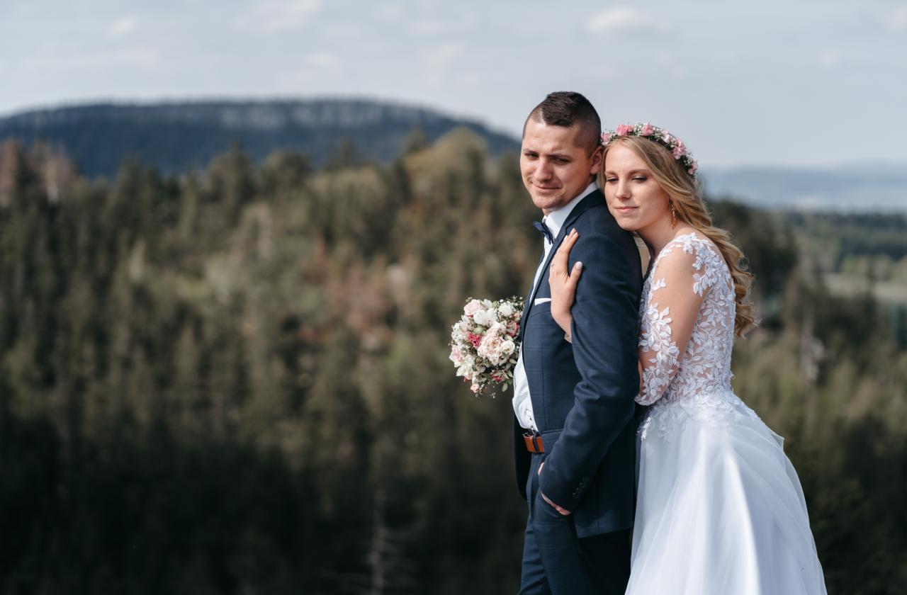 Para młoda w eleganckich strojach pozuje na tle górskiego krajobrazu; panna młoda w wianku z kwiatów opiera głowę o ramię pana młodego, który trzyma bukiet.