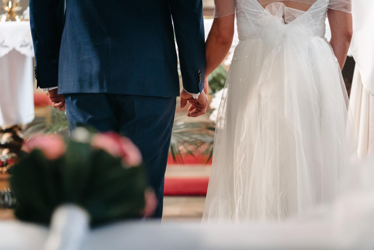 Para młoda trzymająca się za ręce podczas ceremonii ślubnej; widok z tyłu, on w granatowym garniturze, ona w białej sukni ślubnej z kokardą na plecach, w tle ołtarz i czerwony dywan.