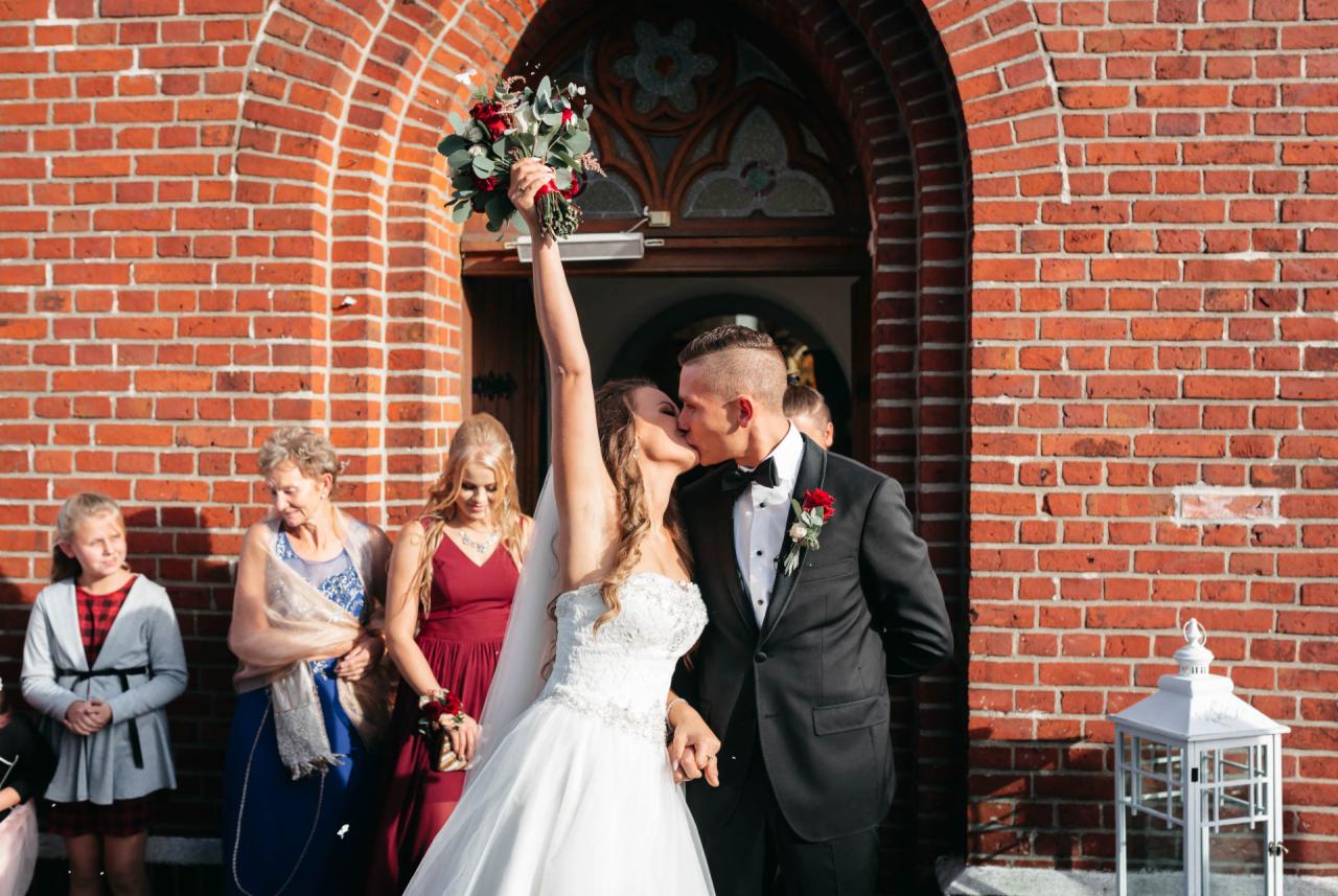 Pocałunek nowożeńców przed ceglanym kościołem, panna młoda z podniesionym bukietem kwiatów, goście weselni w tle.