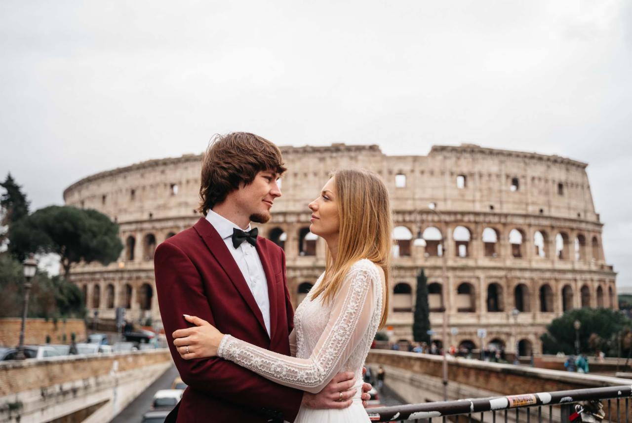 Para młoda w objęciach na tle rzymskiego Koloseum, on w bordowym smokingu z muchą, ona w białej sukni z koronkowymi rękawami, oboje patrzą na siebie z uśmiechem.
