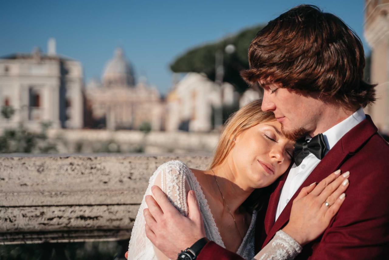 Para młoda w objęciach na tle rzymskiej architektury, panna młoda w koronkowej sukni z zamkniętymi oczami, pan młody w bordowym garniturze z muchą, delikatne światło słoneczne.