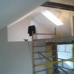 Poddasze w trakcie prac wykończeniowych: ściany po gładziowaniu, widoczne rusztowanie, oświetlenie robocze i czarna kurtka wisząca na ścianie.