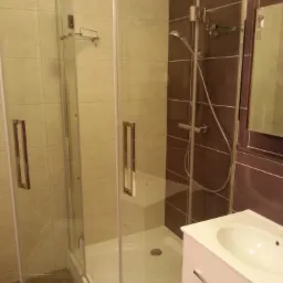 Nowoczesna kabina prysznicowa narożna z białym brodzikiem i umywalką na tle beżowo-brązowych płytek, widok z bliska na wykończoną łazienkę.