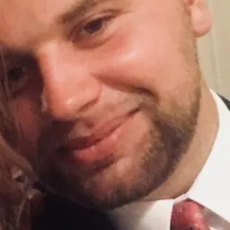 Portret uśmiechniętego mężczyzny z krótką fryzurą i brodą, ubranego w garnitur i wzorzysty krawat, uchwyconego w zbliżeniu.