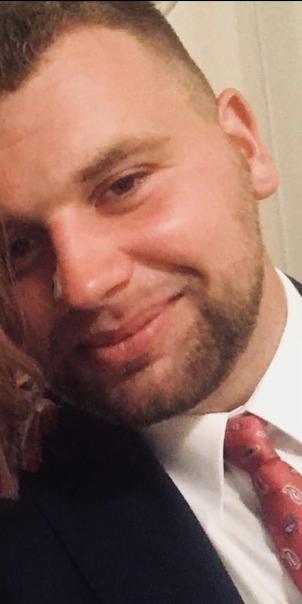 Portret uśmiechniętego mężczyzny z krótką fryzurą i brodą, ubranego w garnitur i wzorzysty krawat, uchwyconego w zbliżeniu.