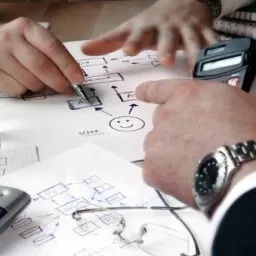 Ręce nad schematem blokowym rysowanym na papierze, z kalkulatorem, szklanką wody i telefonem na biurku, prawdopodobnie podczas sesji planowania biznesowego.