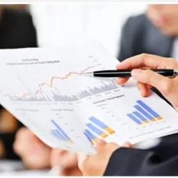 Osoba w garniturze analizuje raport z wykresami i danymi finansowymi, wskazując na rosnącą linię na wykresie za pomocą czarnego długopisu.