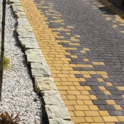 Ułożona kostka brukowa w dwóch kolorach, tworząca wzór. Obrzeże z kamienia naturalnego i żwirowe podsypanie. Widok z góry, ukośny kadr.