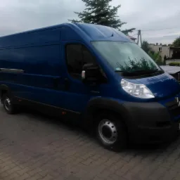 Niebieski furgon Citroen Jumper na kostce brukowej, zaparkowany pod pochmurnym niebem. Widoczne detale karoserii i opon, zarys budynku w tle.