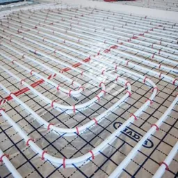 Białe rury ogrzewania podłogowego ułożone na folii z nadrukowaną siatką, spięte czerwonymi uchwytami. Widok z góry, instalacja przed wylewką.