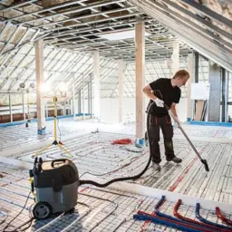 Montaż ogrzewania podłogowego w surowym wnętrzu poddasza, widoczny pracownik z odkurzaczem przemysłowym i lampa na statywie. Instalacja w trakcie czyszczenia.