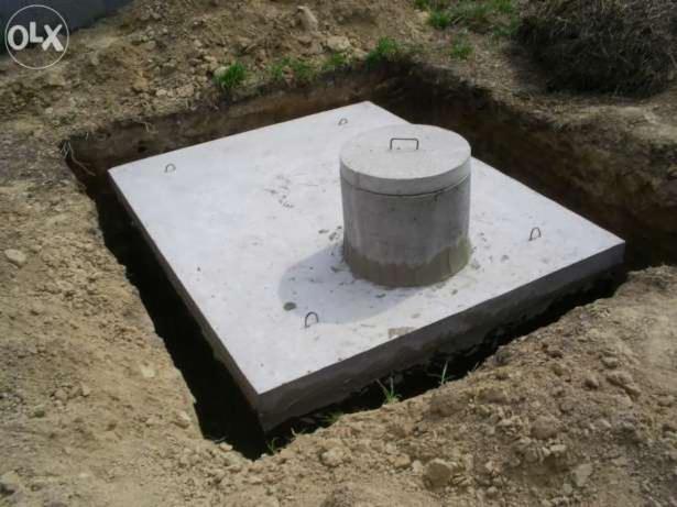 Betonowa płyta szamba z pokrywą w wykopie, widoczne uchwyty montażowe, ziemia wokół, świeży wykop, minimalizm, surowy beton.