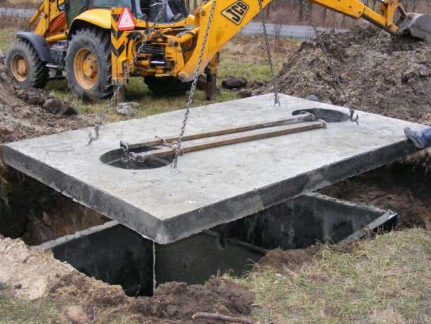 Żółta koparka JCB opuszcza betonową płytę z otworem na szambo do wykopu w ziemi, na łańcuchach. Widoczne brązowe belki. Praca na zewnątrz.