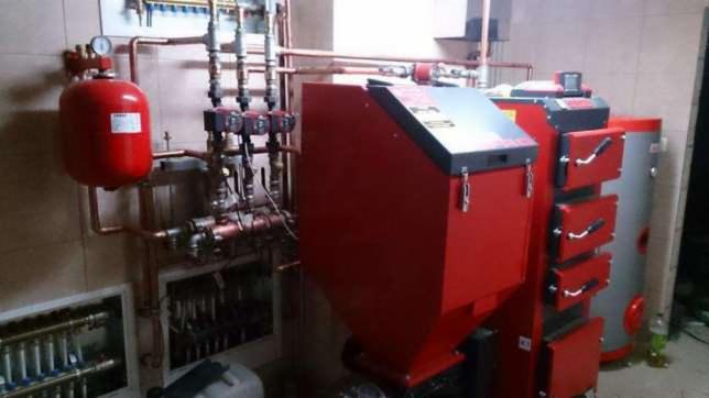Czerwony piec CO z zasobnikiem, rurami miedzianymi i osprzętem w kotłowni. Widoczne manometry i zawory. Instalacja grzewcza w pomieszczeniu technicznym.