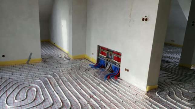 Rozłożona instalacja ogrzewania podłogowego, rury ułożone w pętle na siatce, widoczne przyłącza do rozdzielacza, surowy stan ścian w jasnym wnętrzu.