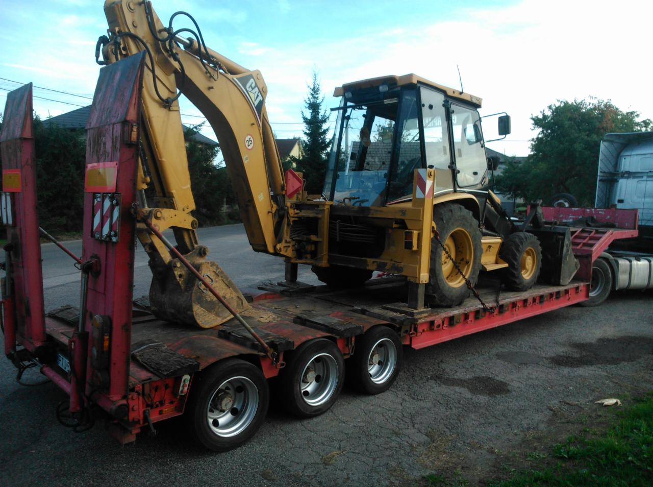 Żółta koparko-ładowarka CAT na niskopodwoziowej platformie transportowej z czerwoną ramą, widok z boku. Na drugim planie drzewa i fragment białej ciężarówki.